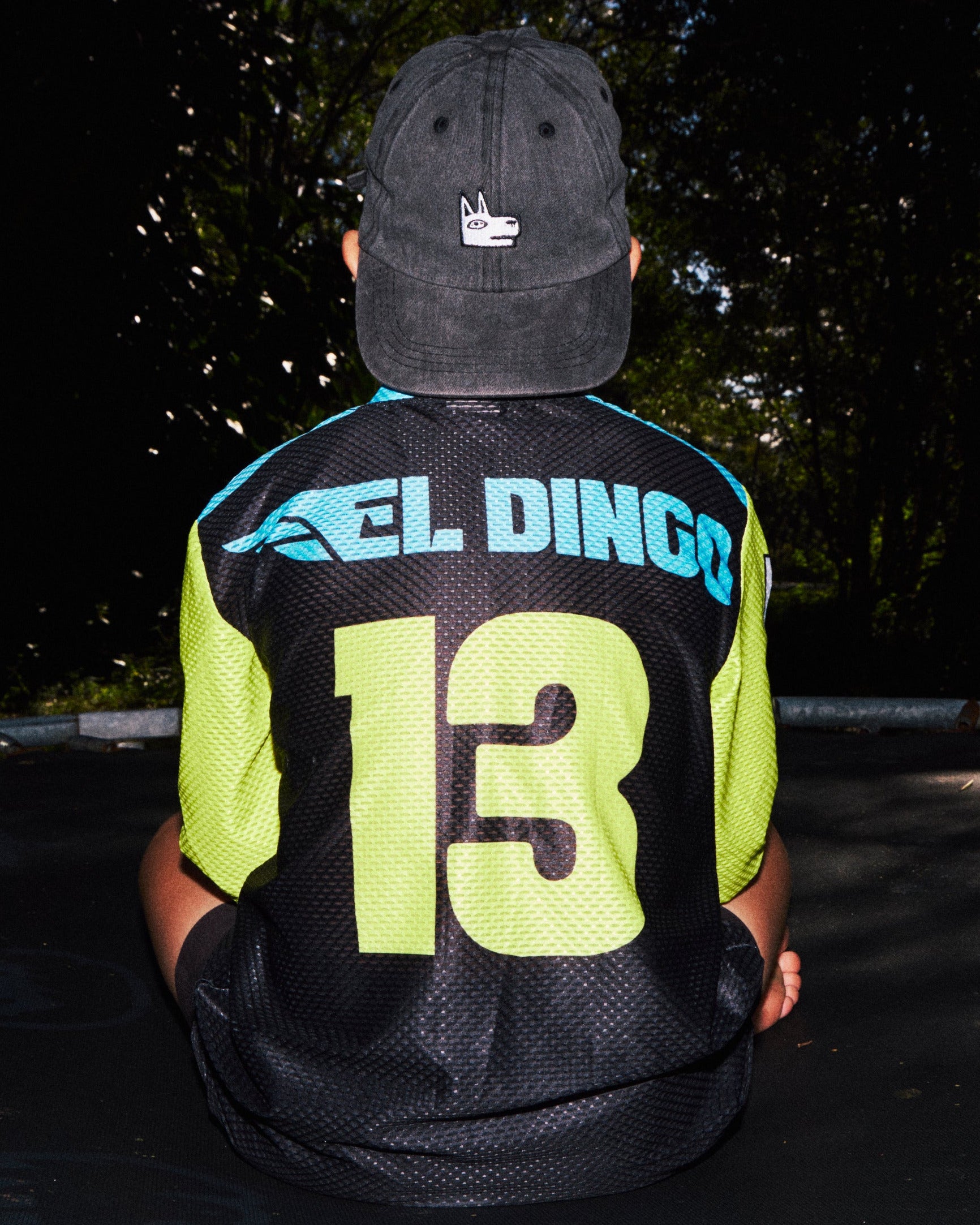 A person sitting on a trampoline outdoors, viewed from behind, wearing a black and neon yellow jersey with the name 'EL DINGO' and the number '13' on the back, along with a dark baseball cap.