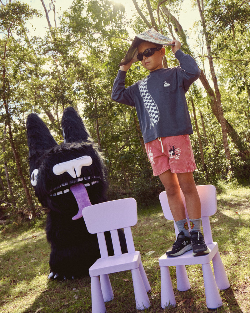 A child wearing a navy graphic sweatshirt, pink shorts, and a white hat stands on a small white plastic chair in a wooded outdoor setting next to our el dingo mascot costume.