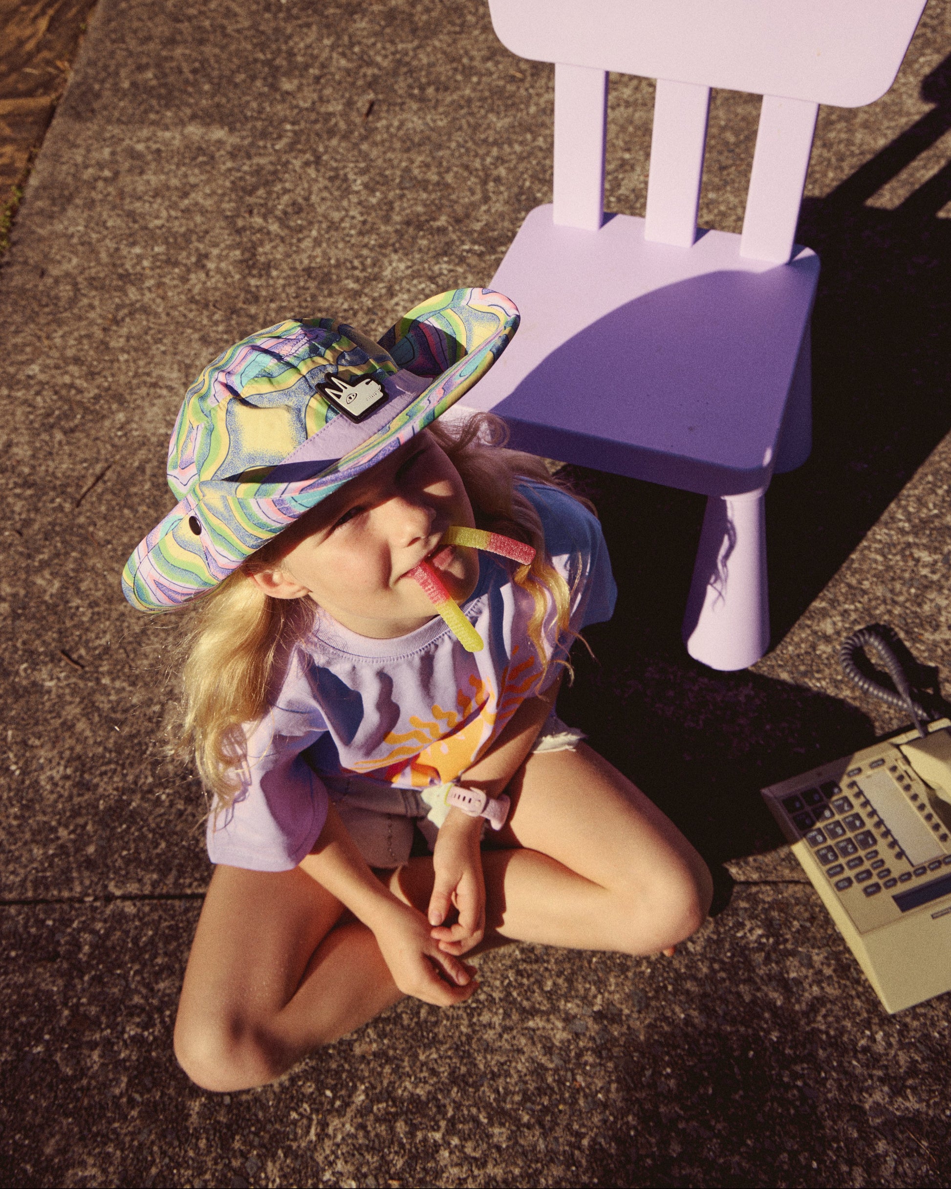A young child wearing a colorful patterned sun hat and a purple shirt sits on a concrete patio next to a matching purple plastic chair.
