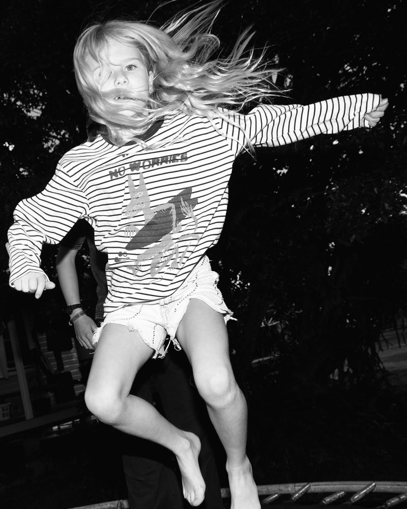 A black and white action photograph of a young girl jumping energetically on a trampoline, wearing a horizontally striped long-sleeve t-shirt and denim shorts.