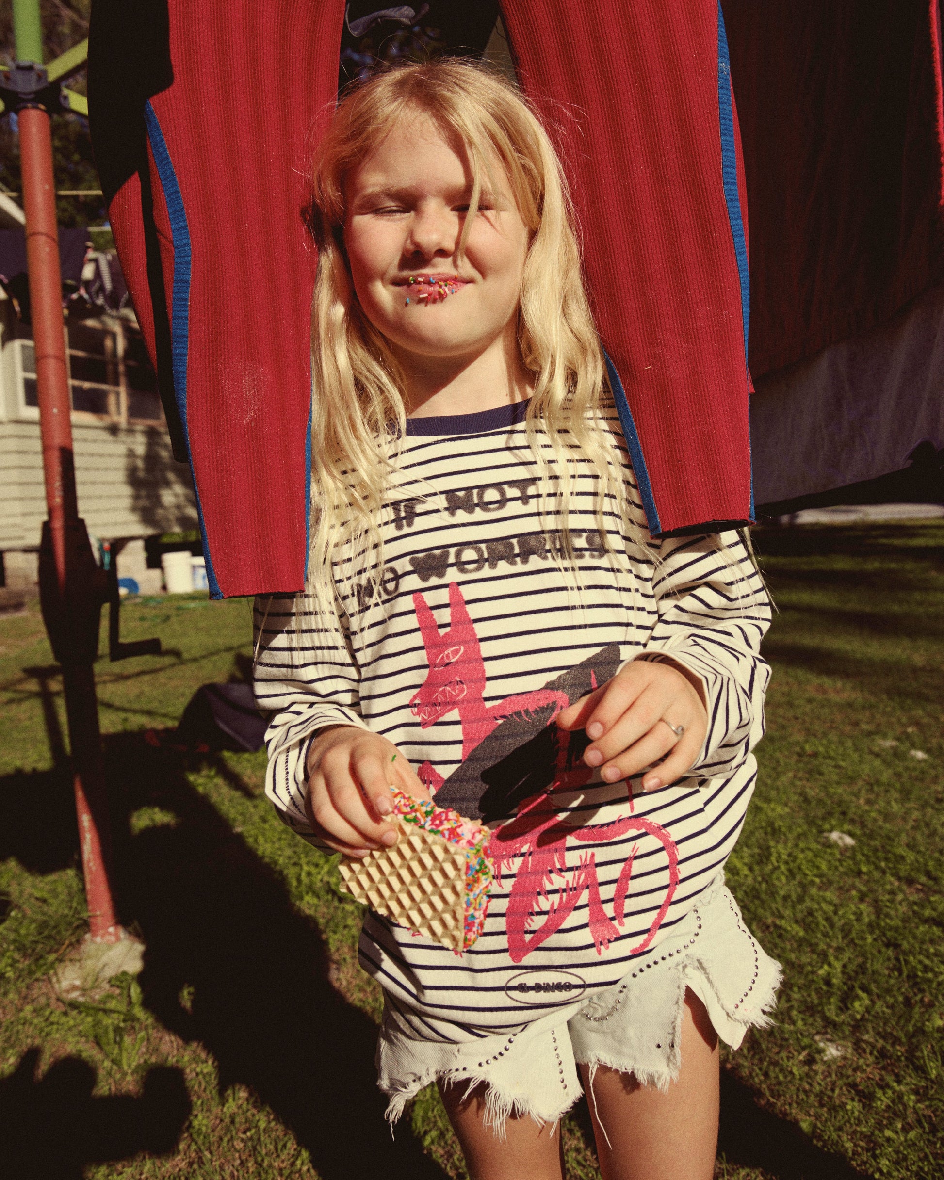 A young blonde girl stands outdoors on grass wearing a black-and-white striped long-sleeve shirt with a pink graphic and denim shorts. She is holding a small woven basket and a black object in front of a red and blue striped hanging garment.