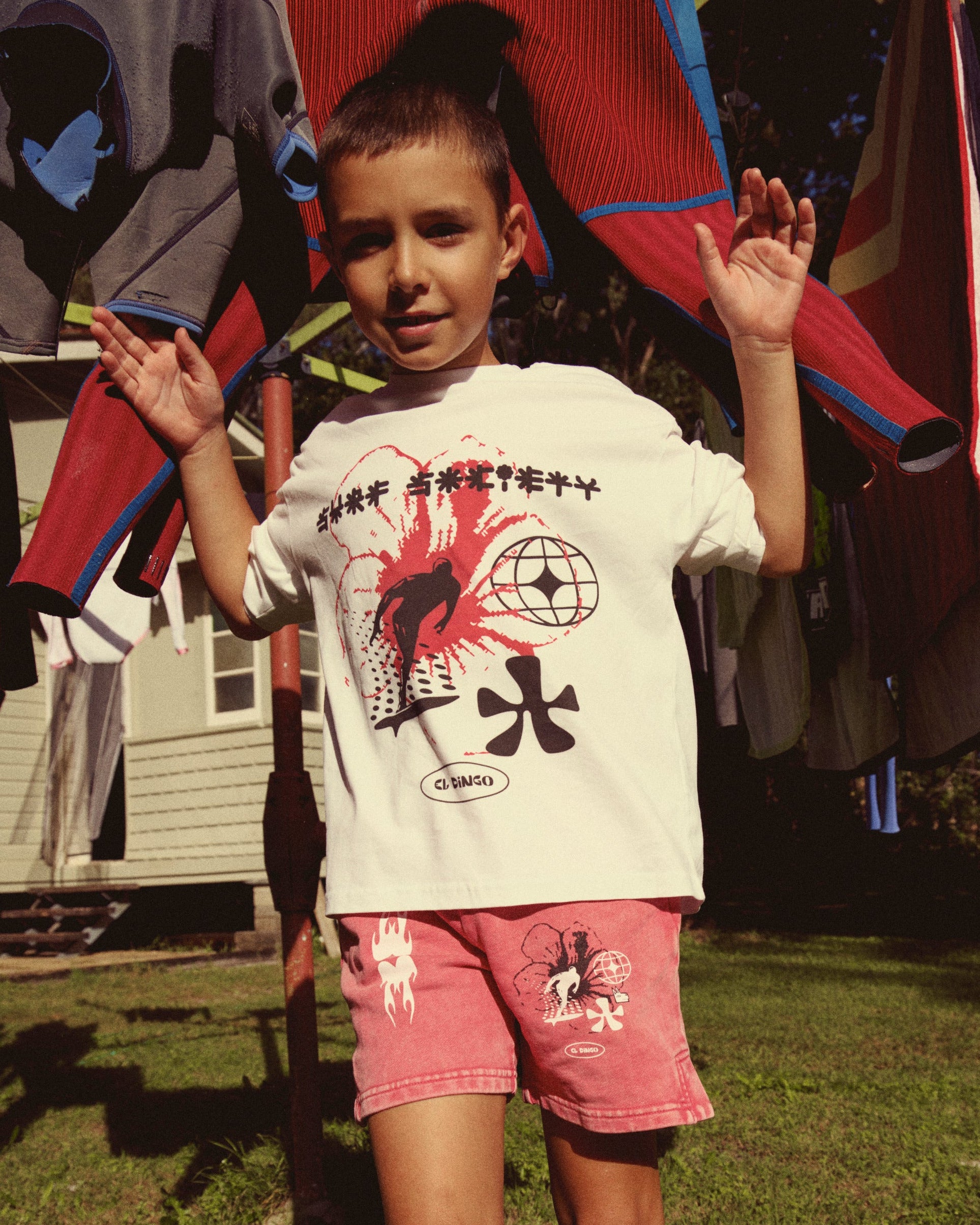 A young boy stands on a grassy lawn wearing a white graphic t-shirt and pink shorts, with colorful sports jerseys hanging on a clothesline in the background.