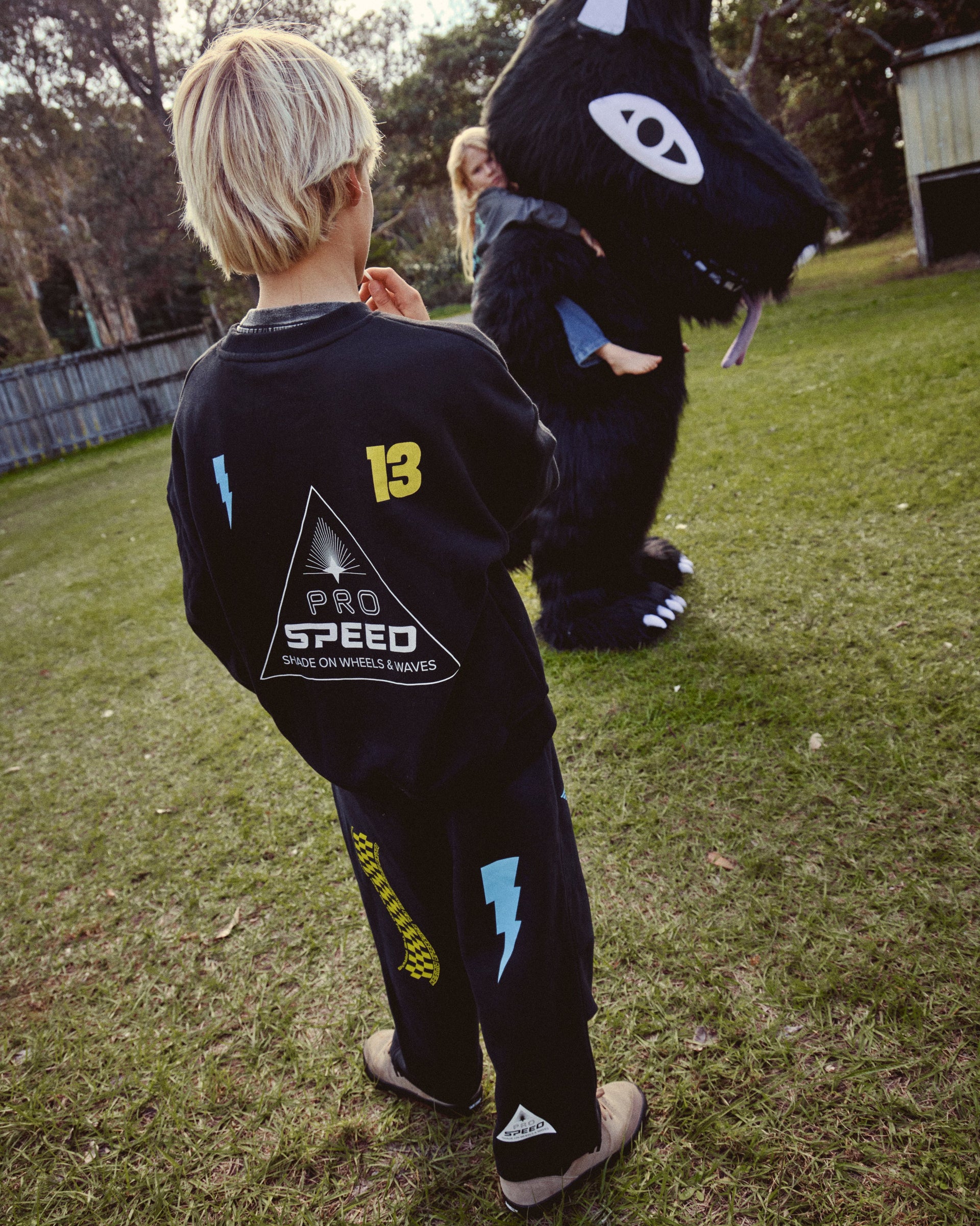 A child wearing a black Pro Speed long-sleeve shirt and matching pants stands on a grassy lawn, facing a person in a large black wolf mascot costume. The shirt features a yellow '13' and a white triangular logo with 'PRO SPEED' text.