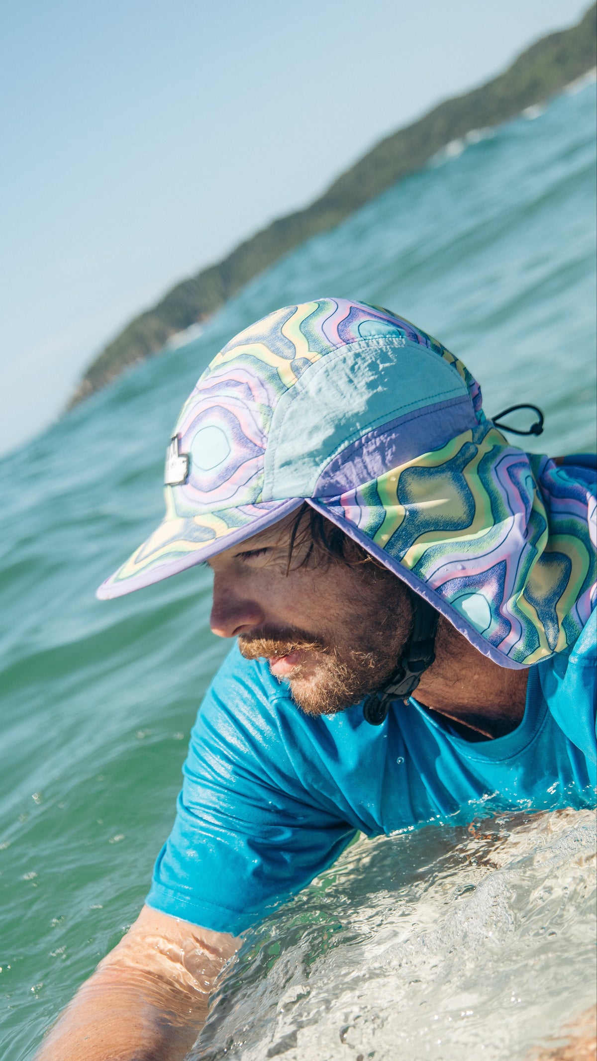 A man in a blue shirt wearing a colorful patterned sun hat with a neck shield, leaning over the edge of a wave in the ocean.
