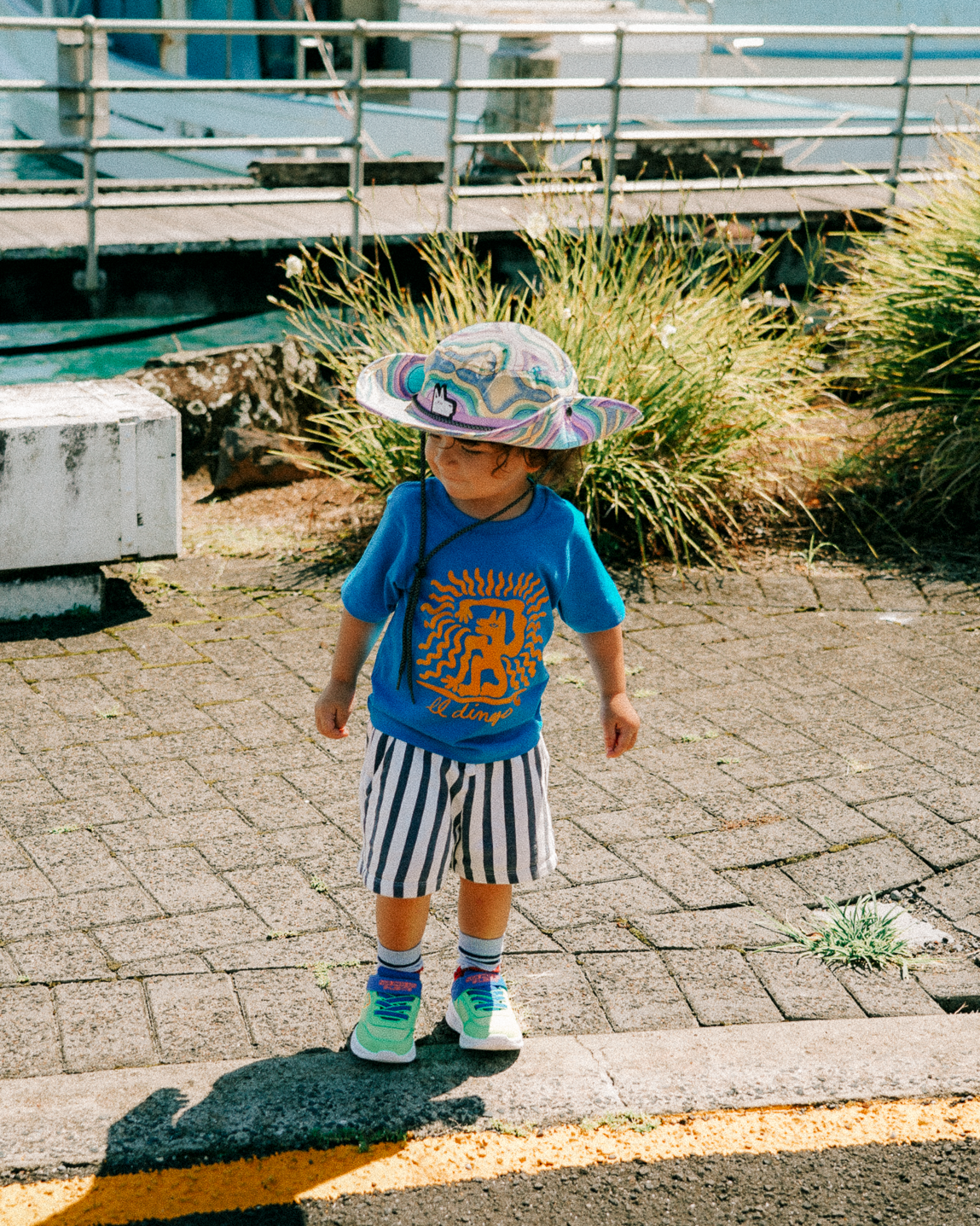 Child wearing a  colourful hat, blue shirt and striped shorts standing on a dock with boats in the background.