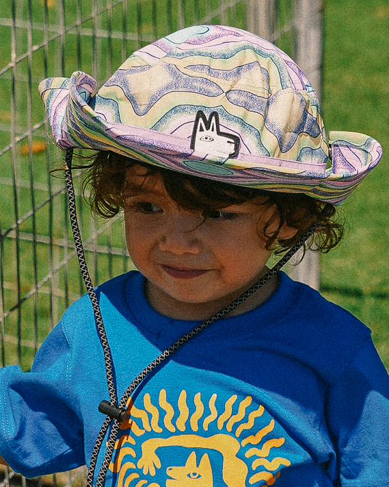 Child wearing a colourful upf 50 sun hat blue shirt with a yellow design, striped shorts, and colorful shoes standing next to a metal fence.