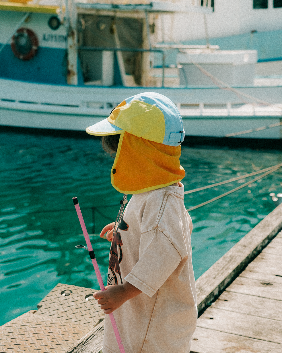 Child wearing a colorful hat and yellow raincoat by a dock with boats in the background