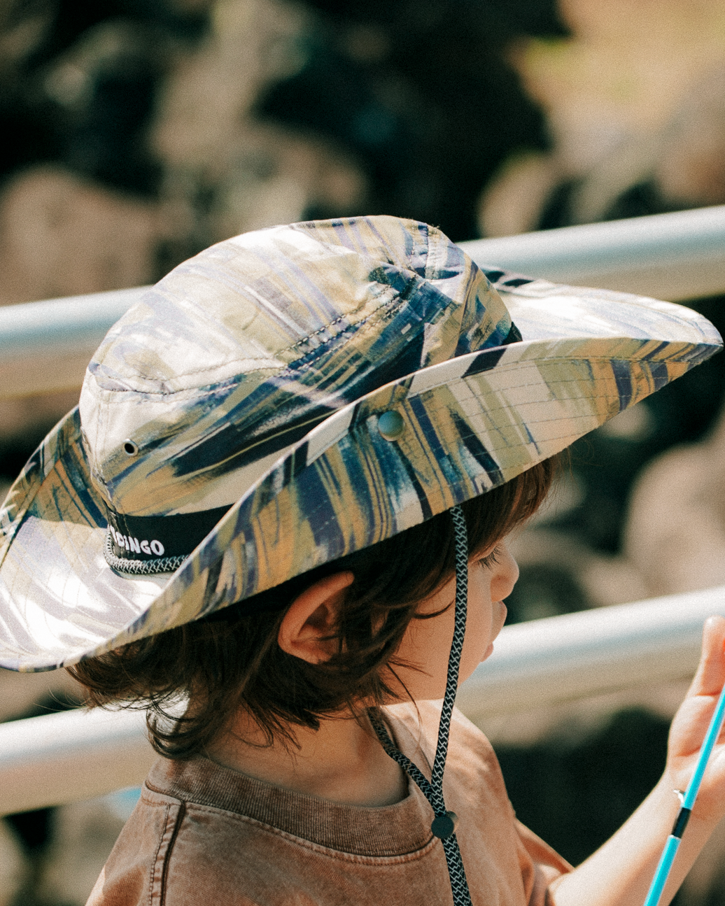 Child wearing a patterned sun hat and brown shirt with an orange design, holding a blue stick.