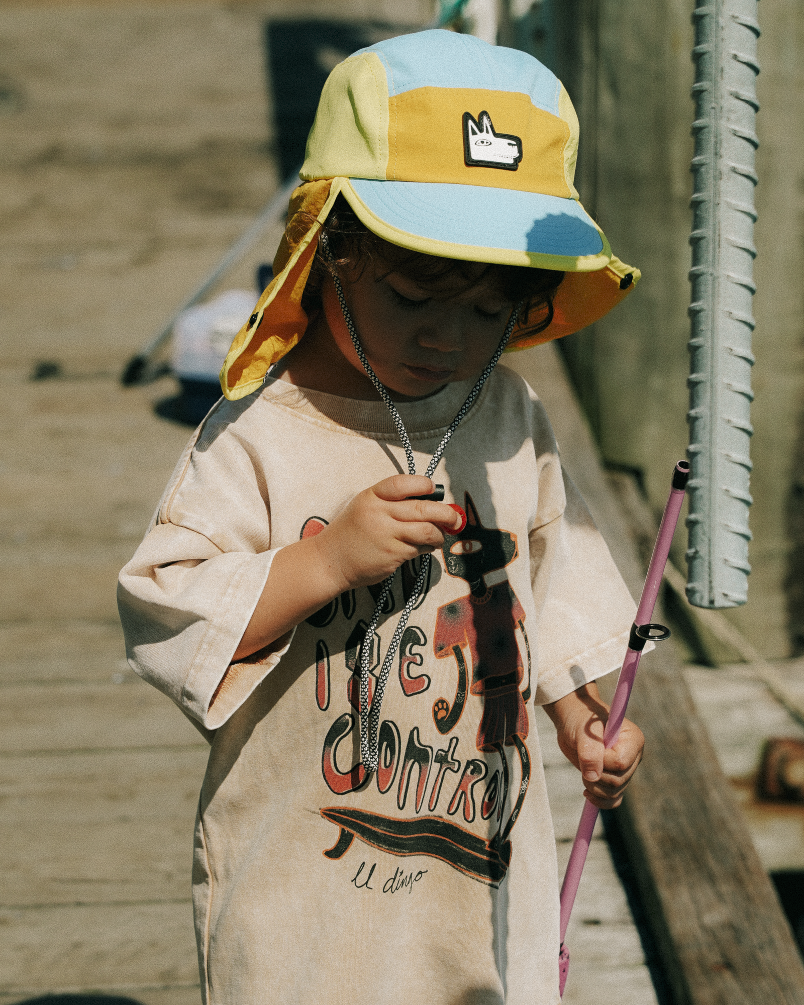 Child wearing a colorful surf hat and holding a pink object, standing on a wooden deck.
