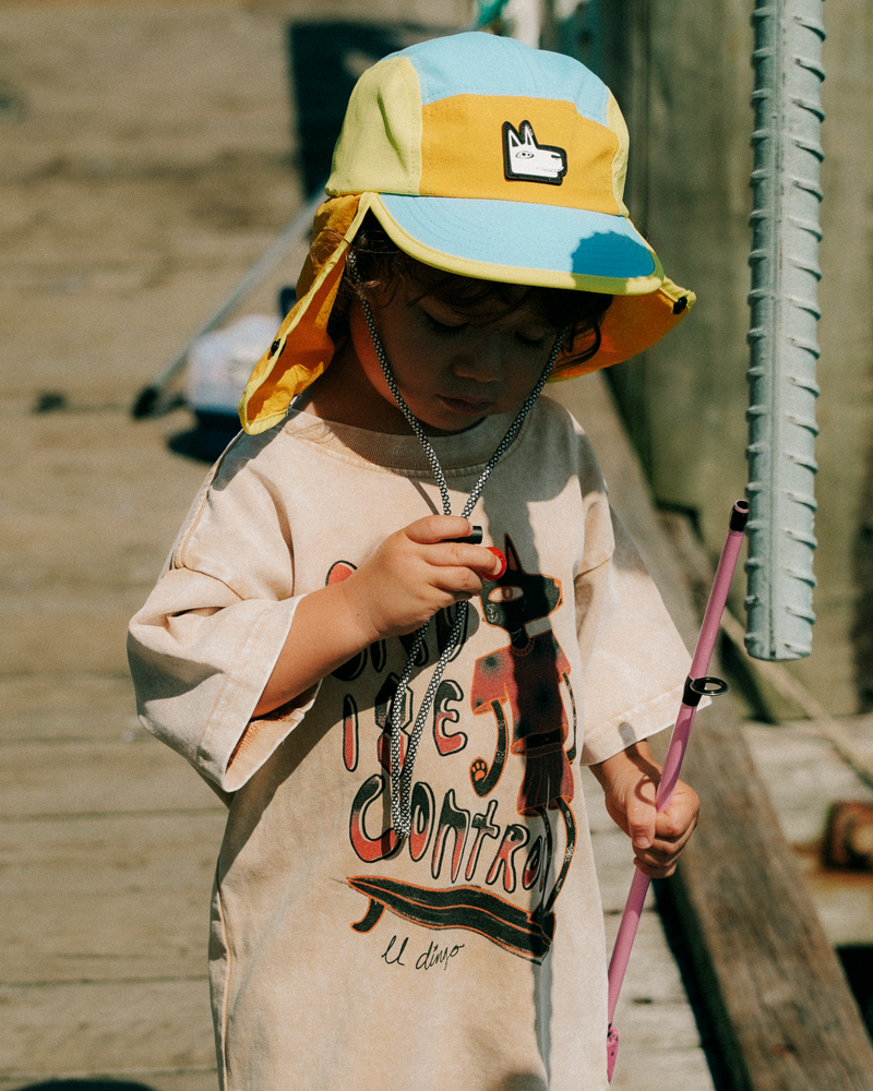 Child wearing a colorful surf hat and holding a pink object, standing on a wooden deck.